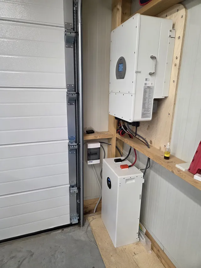 Deye inverter on a timber board in a garage beside the garage door, with one battery module below.
