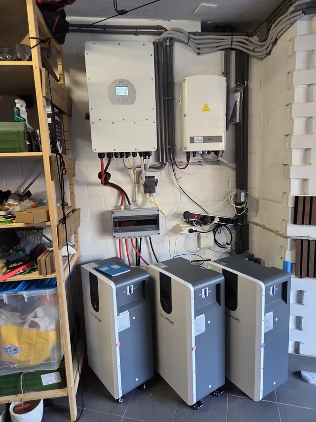 Deye and SolarEdge inverters on a white-painted brick wall, with three grey Exliporc battery cabinets on castors below.